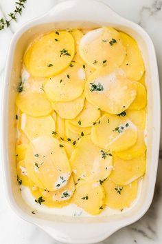 a casserole dish filled with potatoes and cheese on a marble countertop next to herbs