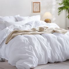 an unmade bed with white linens and blankets on it in a room next to a potted plant