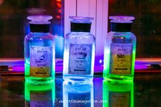 three bottles are sitting on a table with colored lights in the window sill behind them