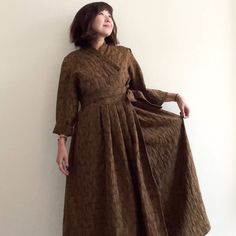 a woman standing in front of a white wall wearing a brown dress and black shoes