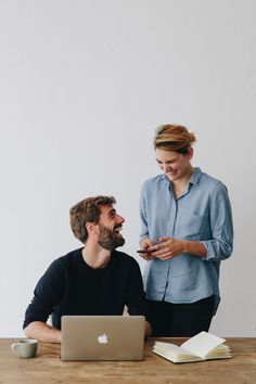 a man and woman sitting at a table looking at something on their cell phone, with an open laptop in front of them