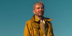 a man with blonde hair wearing a yellow jacket and looking at the camera while standing in front of a blue sky