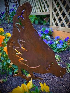 a metal rooster statue sitting in the middle of some flowers and plants with blue, yellow, orange, and red flowers around it