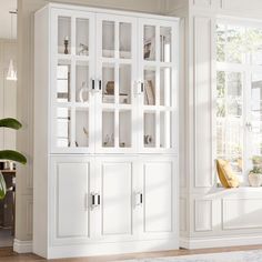 a large white bookcase with glass doors in a living room next to a window