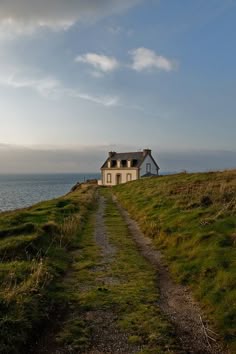 a house sitting on top of a lush green hillside next to the ocean in front of a cloudy blue sky