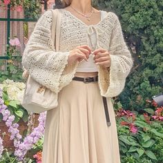 a woman standing in front of flowers wearing a white sweater and beige pleated skirt