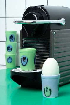 there are cups and an egg on the counter next to a coffee machine with three cups in front of it