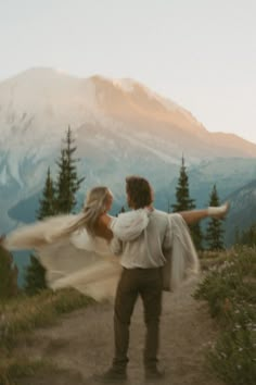 a man and woman holding each other on top of a hill with mountains in the background