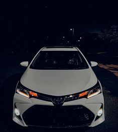 the front end of a white car parked in a parking lot at night with its lights on