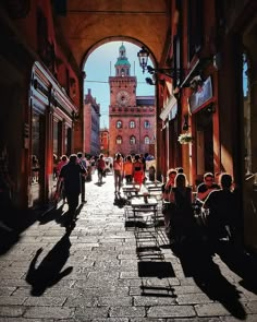 people are sitting at tables in an alleyway with buildings and a clock tower behind them