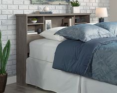 a bed with blue sheets and pillows in front of a white brick wall next to a potted plant