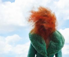 a woman with red hair standing in front of a blue sky wearing a green coat