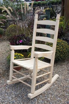 a wooden rocking chair sitting on top of a gravel ground next to bushes and flowers