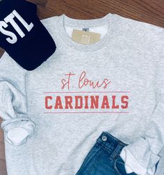 the st louis cardinals sweatshirt and hat are laying on top of a wooden floor next to jeans