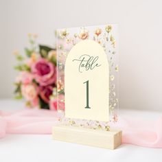 a table number is placed on a wooden stand with flowers in the background and pink ribbon