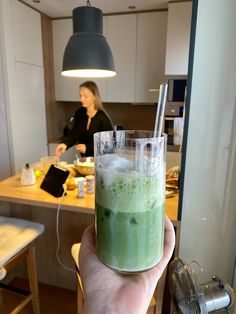 a person holding up a glass with green liquid in it and a woman sitting at the kitchen counter behind them