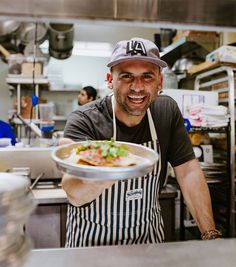 a man in an apron and hat holding a plate with food on it while smiling at the camera