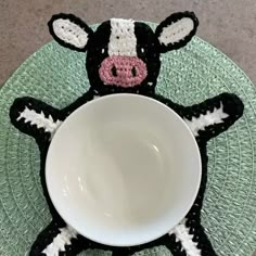 a crocheted cow sitting next to a white bowl on a green place mat