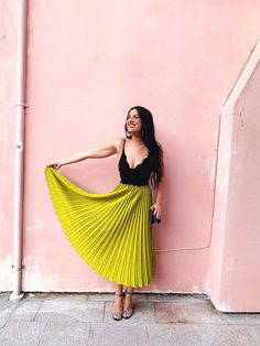 a woman standing against a pink wall wearing a yellow pleaed skirt and black top