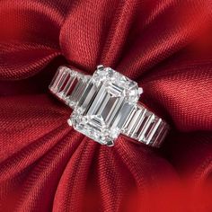 an emerald - cut diamond ring sits atop a red satin fabric, surrounded by three baguets