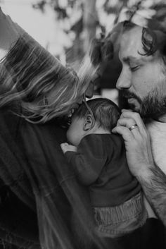 Black and white photo taken through a window or a mother and father hugging their newborn son between them and kissing his head.