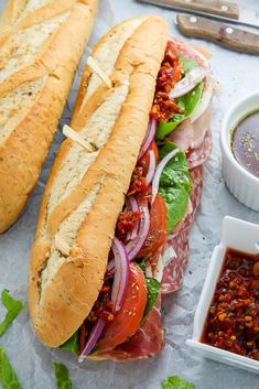 two sub sandwiches sitting on top of a table next to dipping sauces and condiments