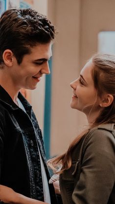 a young man and woman standing next to each other in front of a window looking at each other