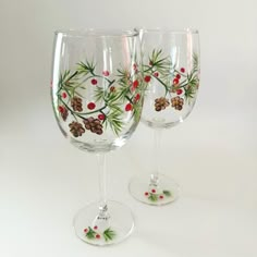 two wine glasses decorated with pine cones and holly berry design on the bottom one glass is clear