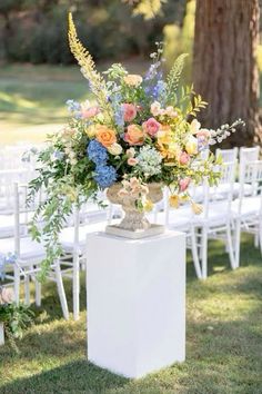 a vase filled with flowers sitting on top of a white pedestal next to chairs in the grass