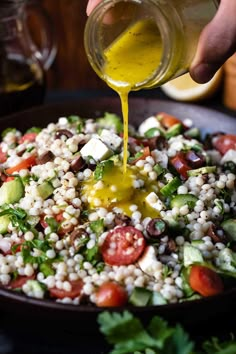 someone pouring dressing onto a salad in a black bowl with tomatoes, cucumbers and other vegetables