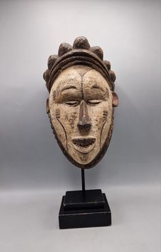 an african mask is displayed on a black stand against a gray background, with the head tilted to the side