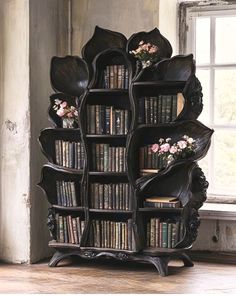 an ornate bookcase with many books and flowers on the top shelf, in front of a window