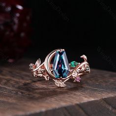 a ring with blue and green stones sitting on top of a wooden table next to a red rose