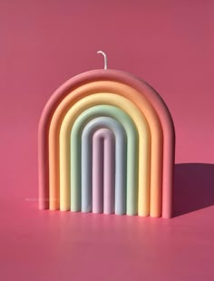 a rainbow shaped candle sitting on top of a pink surface with a single lit candle in the middle