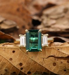 an emerald and diamond ring sitting on top of a leaf covered ground with leaves around it