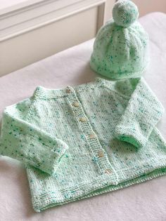 a green knitted sweater and hat sitting on top of a white bed next to a window