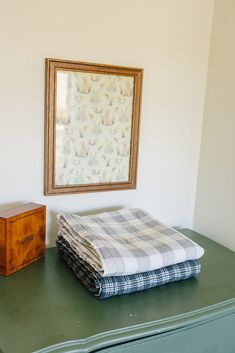 two folded blankets on top of a green dresser next to a framed painting and wooden drawer