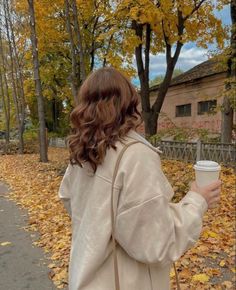 a woman is walking down the street holding a cup in her hand and looking at leaves on the ground