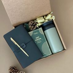 an open box containing a coffee cup, travel mug and pine cones with a pen