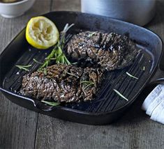 two steaks are cooking on a grill with lemon wedges and herbs in the foreground