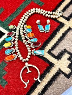 This stunning multi stone squash blossom necklace is crafted by Navajo artist Selena Warner. It is handcrafted with sterling silver and features a stunning array of stones from turquoise to coral, showcasing her expertise in traditional Native American design. The necklace is proudly made in the USA.