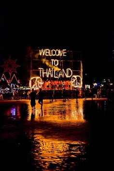 people walking in the rain at night with lights on and welcome to thailand sign lit up