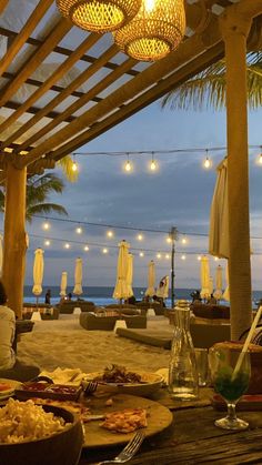 an outdoor dining area on the beach with lights strung over it and people sitting at tables under umbrellas