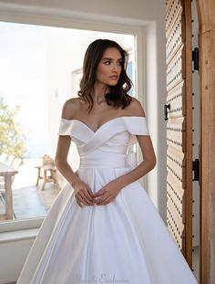 a woman wearing a white wedding dress standing in front of a window with her hands on her hips