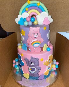 a three tiered cake decorated with teddy bears and rainbows on the top, sitting in a cardboard box