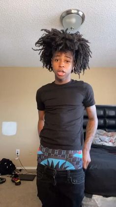 a man with dreadlocks on his head standing in front of a bed and looking at the camera