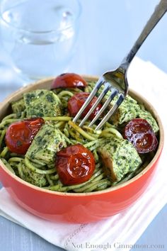 a fork in a red bowl filled with pesto pasta and cherry tomatoes, on top of a white napkin