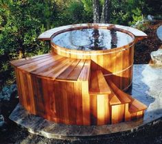 a wooden hot tub sitting on top of a stone walkway next to a lush green forest