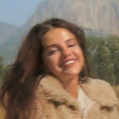 a woman with long brown hair smiling and wearing a fur coat in front of mountains