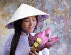 a woman wearing a white hat and holding a pink flower in her right hand while looking at the camera